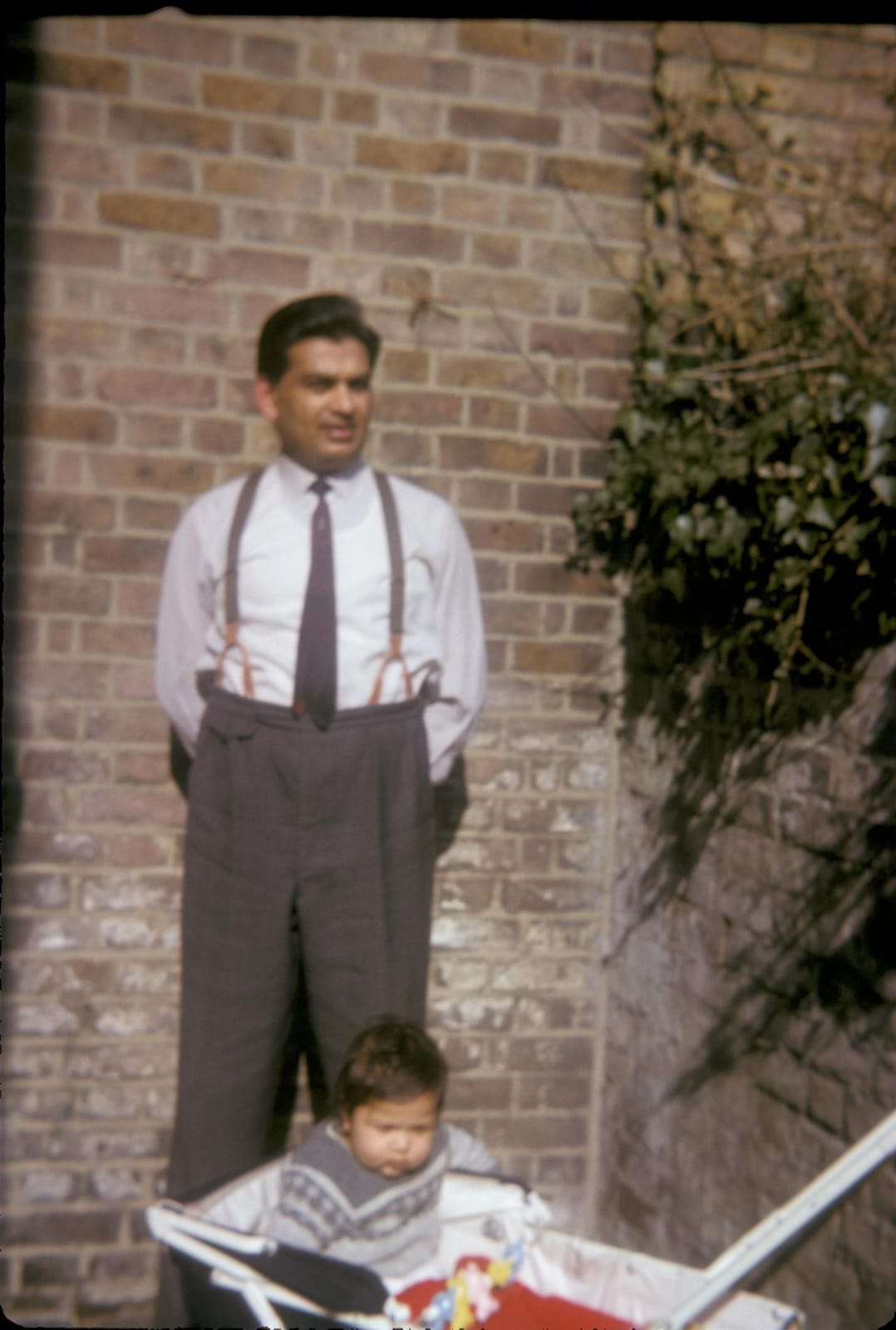 Young Ram Prashad Gupta in shirt and braces by a brick wall in England, a baby in a pram